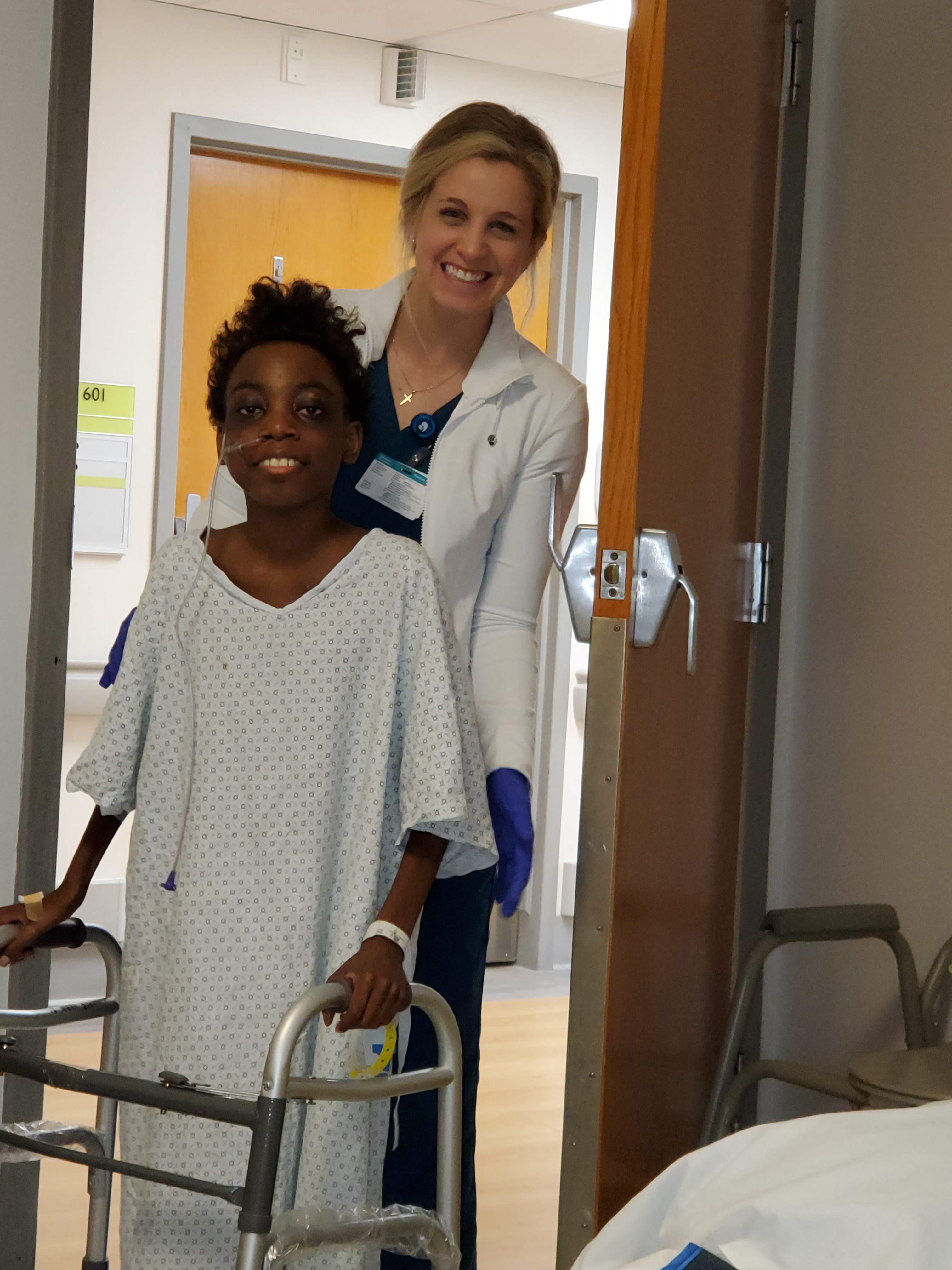Young Abram, wearing a hospital gown, stands with his walker and a Manning Family Children's team member