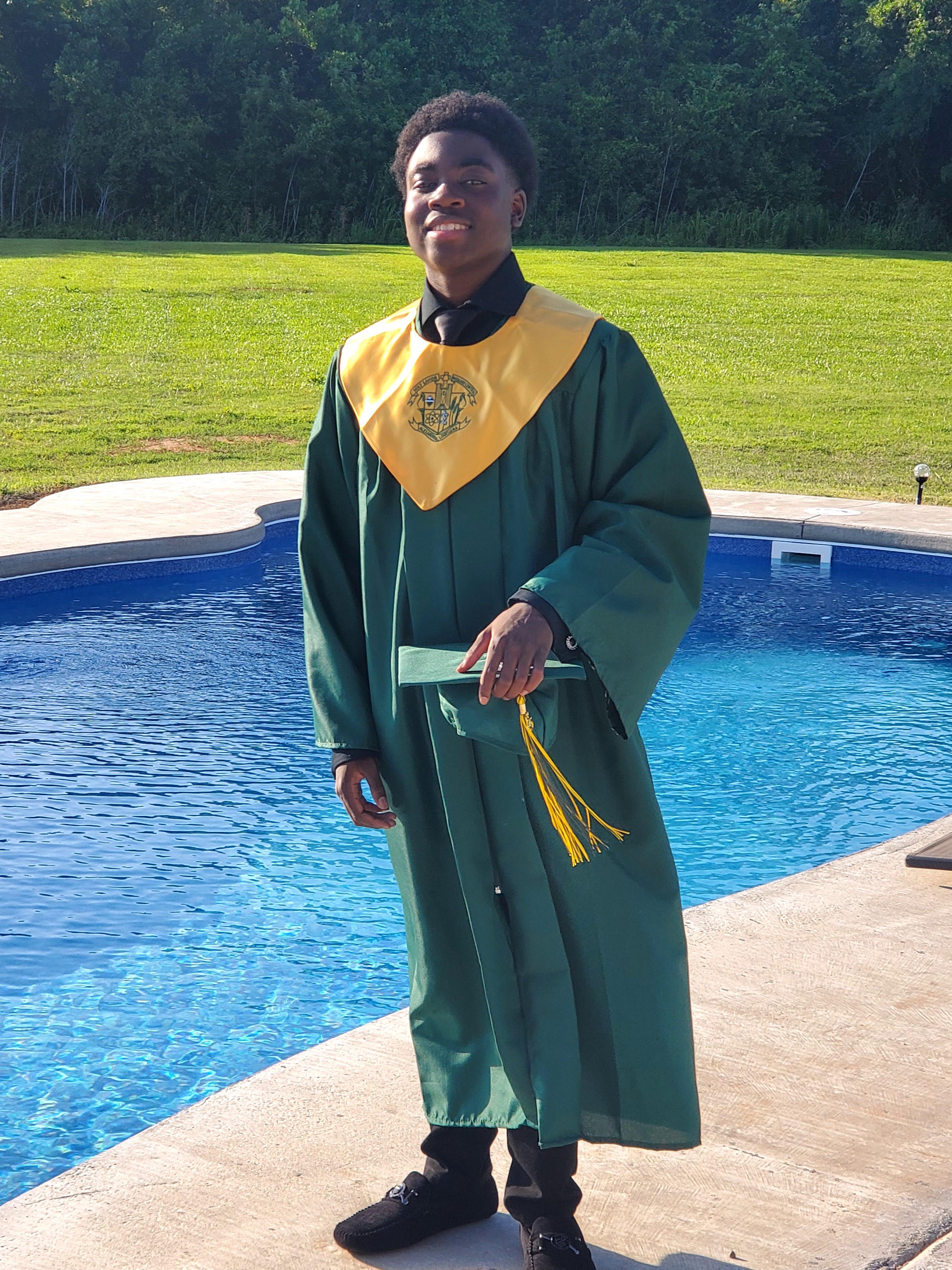Abram stands in front of a backyard pool wearing his graduation gown and holding his graduation cap with a big smile on his face.