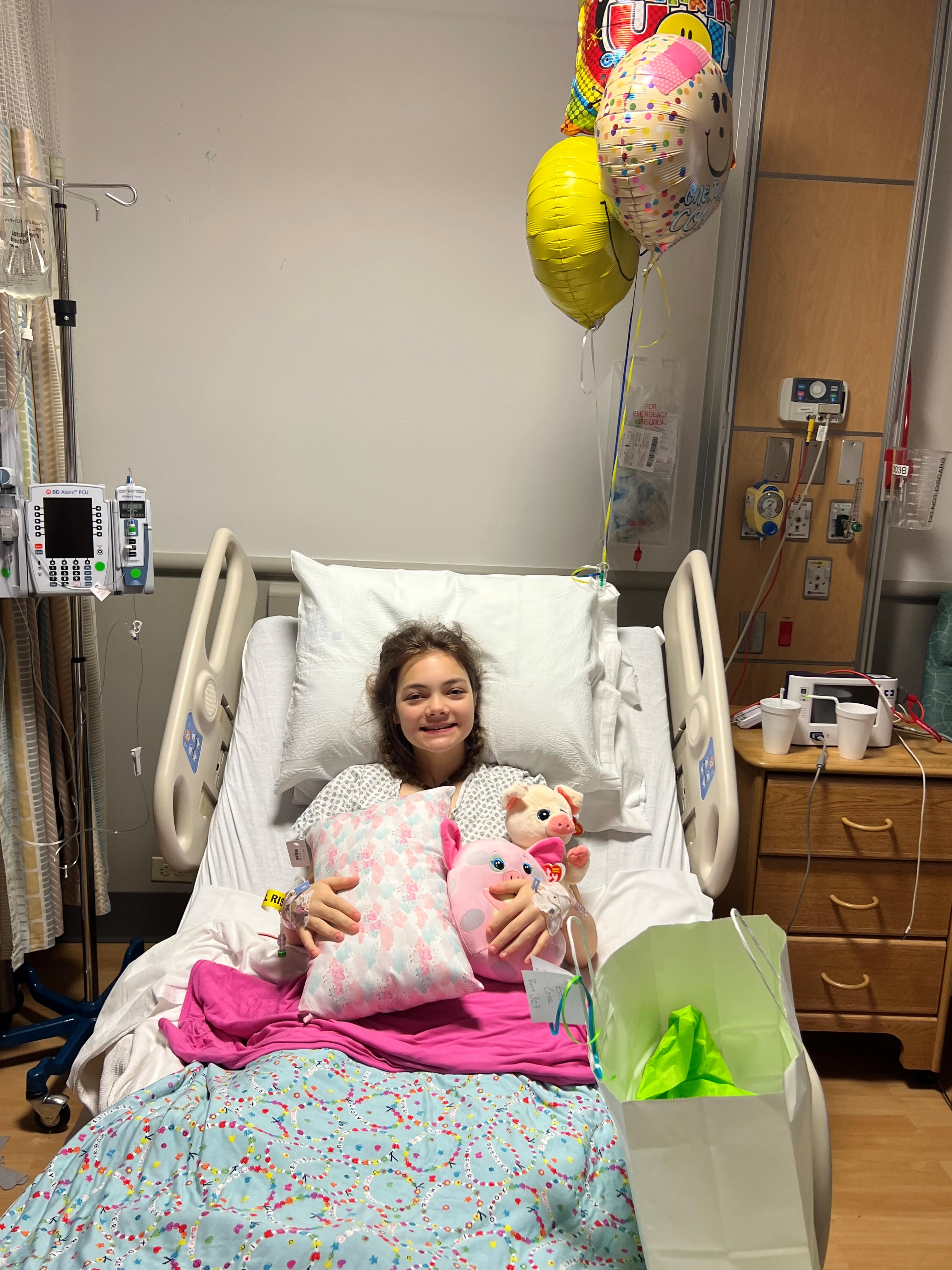 Erin smiles in a hospital bed surrounded by gifts and balloons after a successful hip dysplasia surgery procedure