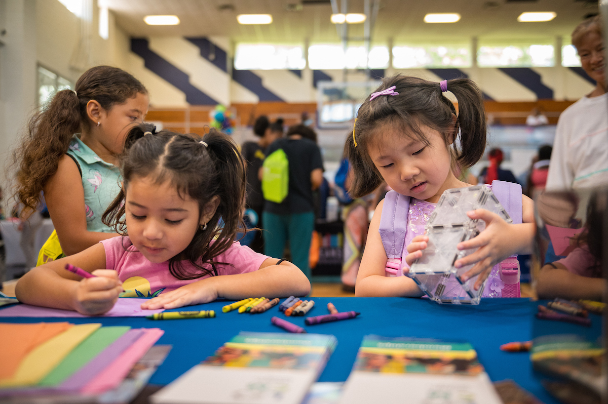 Two young girls sit at a table, coloring and playing with a toy, while other people are in the background.